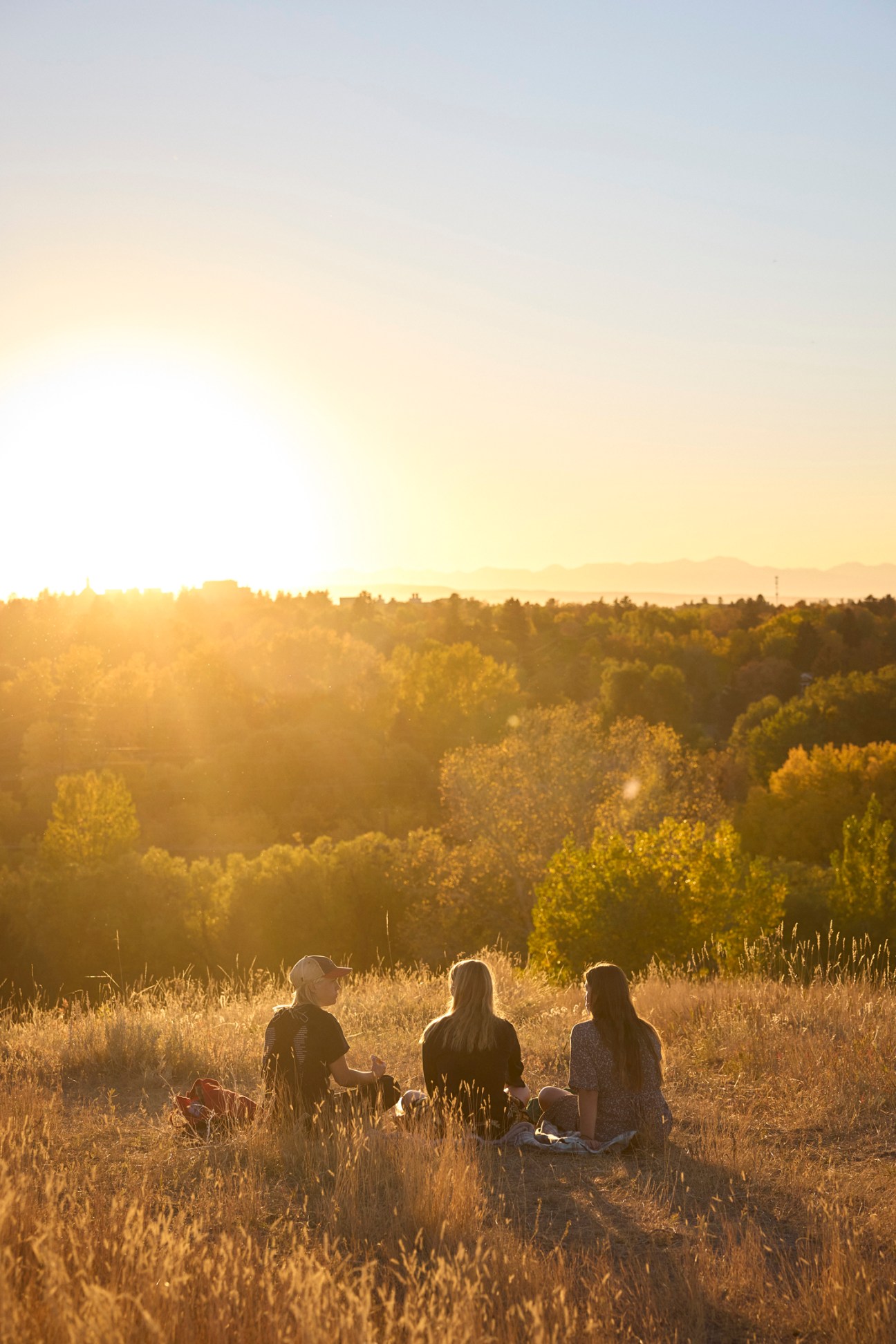 Three people sitting in a grassy field at sunset with forested hills and mountains in the background.