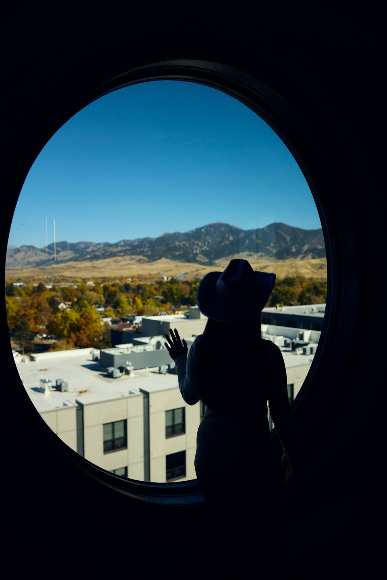 Silhouetted figure in cowboy hat looking out circular window at Bozeman mountains and cityscape.