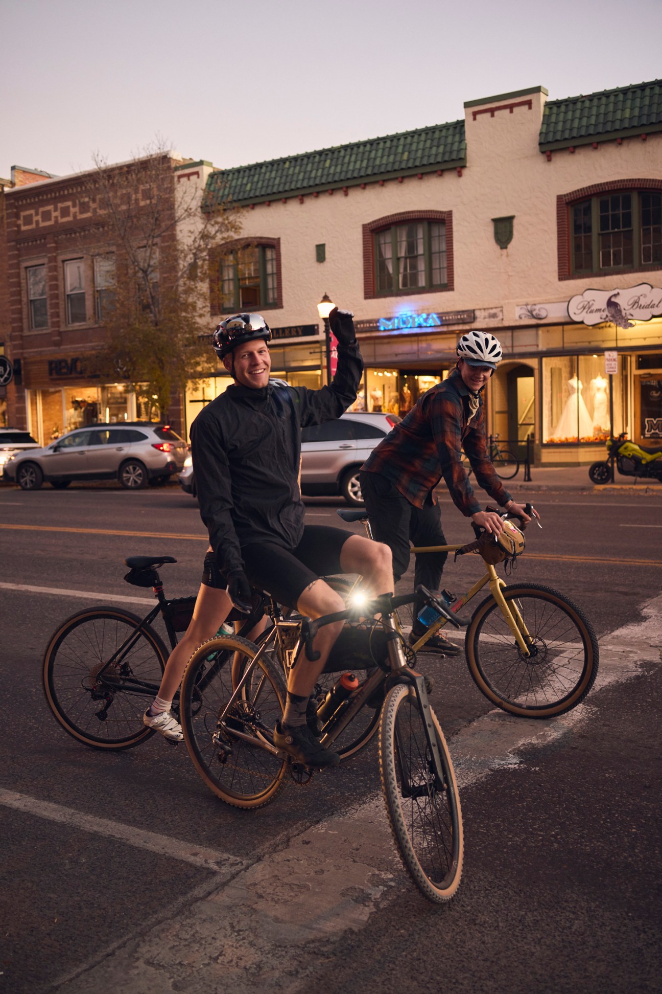 Two cyclists ride bikes on an evening street in Bozeman, Montana, with historic brick buildings and storefronts behind them.