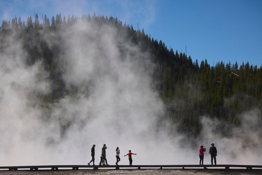 Visitors walk along a wooden boardwalk with geothermal steam rising from hot springs and forested mountains behind in Bozeman, Montana.