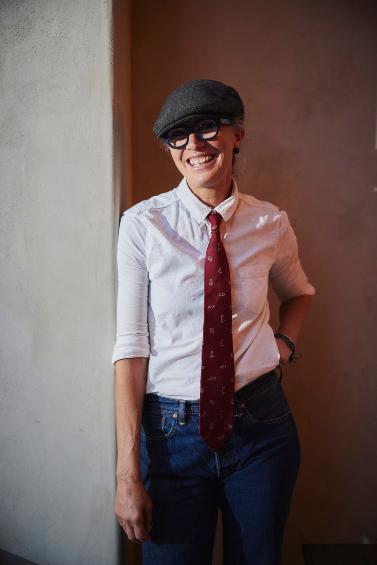 Person wearing a dark cap, glasses, white shirt and burgundy tie smiling against a beige wall.