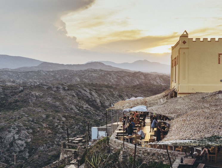 Restaurant terrace with diners overlooking rugged Mediterranean coastline at sunset