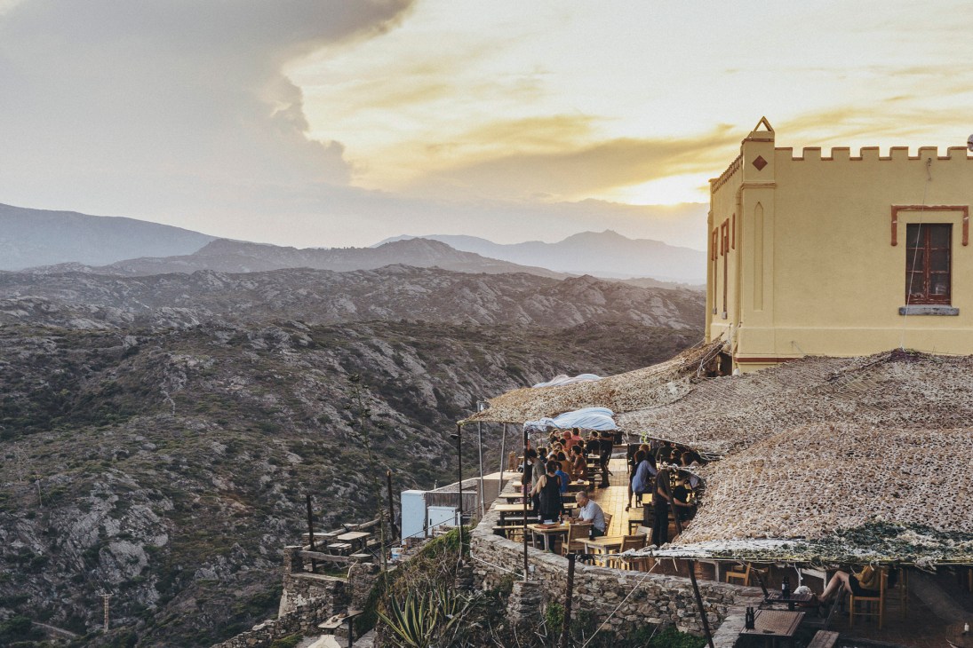 Restaurant terrace with diners overlooking rugged Mediterranean coastline at sunset