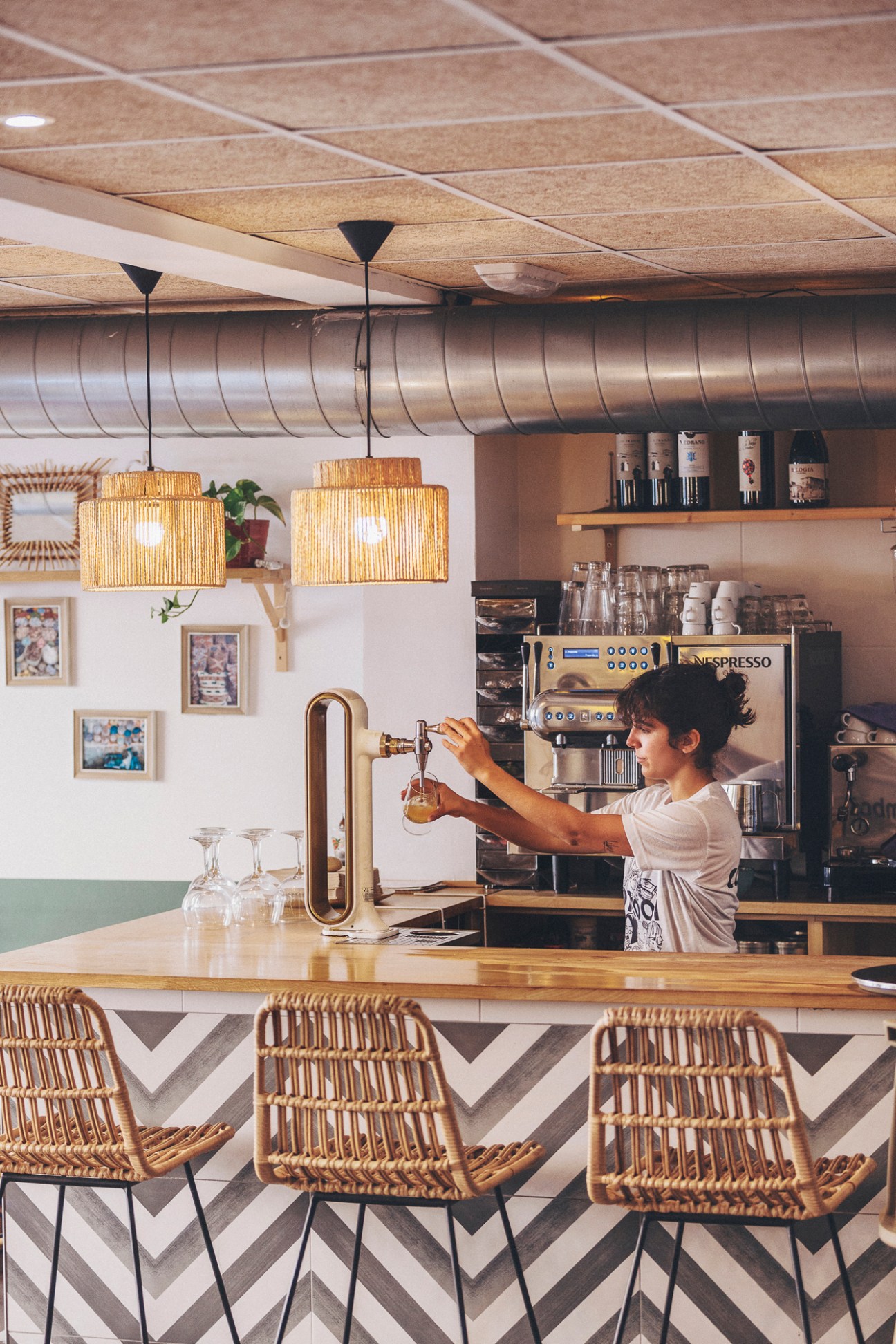 Coffee shop interior with barista preparing drinks behind counter, featuring wicker pendant lights and bar seating