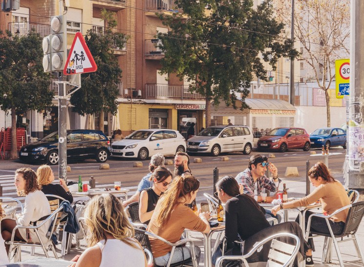 People dining at outdoor cafe terrace with aluminum chairs facing residential buildings and parked cars