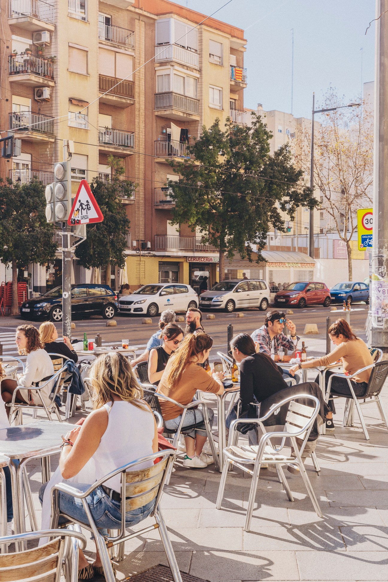 People dining at outdoor cafe terrace with aluminum chairs facing residential buildings and parked cars