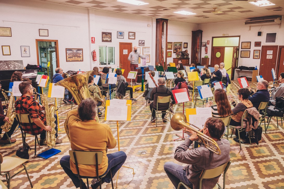 Musicians of all ages seated with brass and wind instruments during orchestra rehearsal in community center