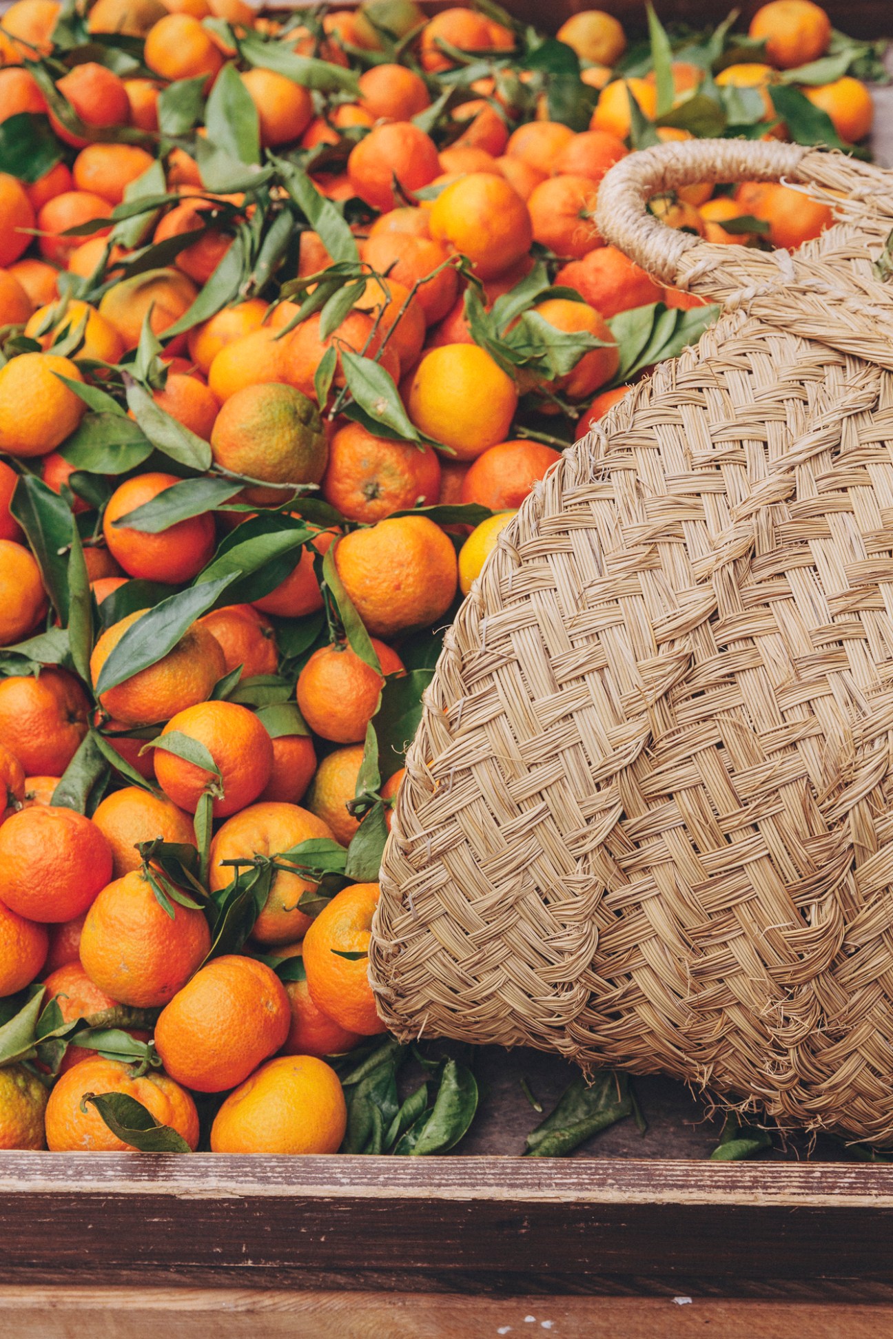 Fresh oranges with green leaves scattered around a woven basket on wooden surface