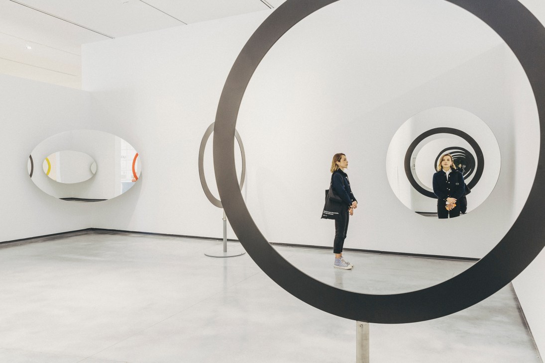 Two visitors view circular mirror installations in a white contemporary art museum gallery space