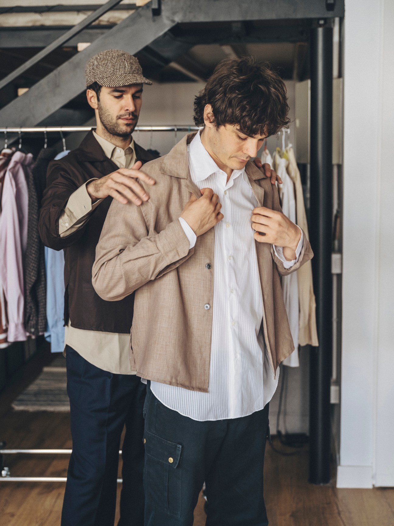 Two men in clothing store, one helping the other try on a beige corduroy jacket over white striped shirt