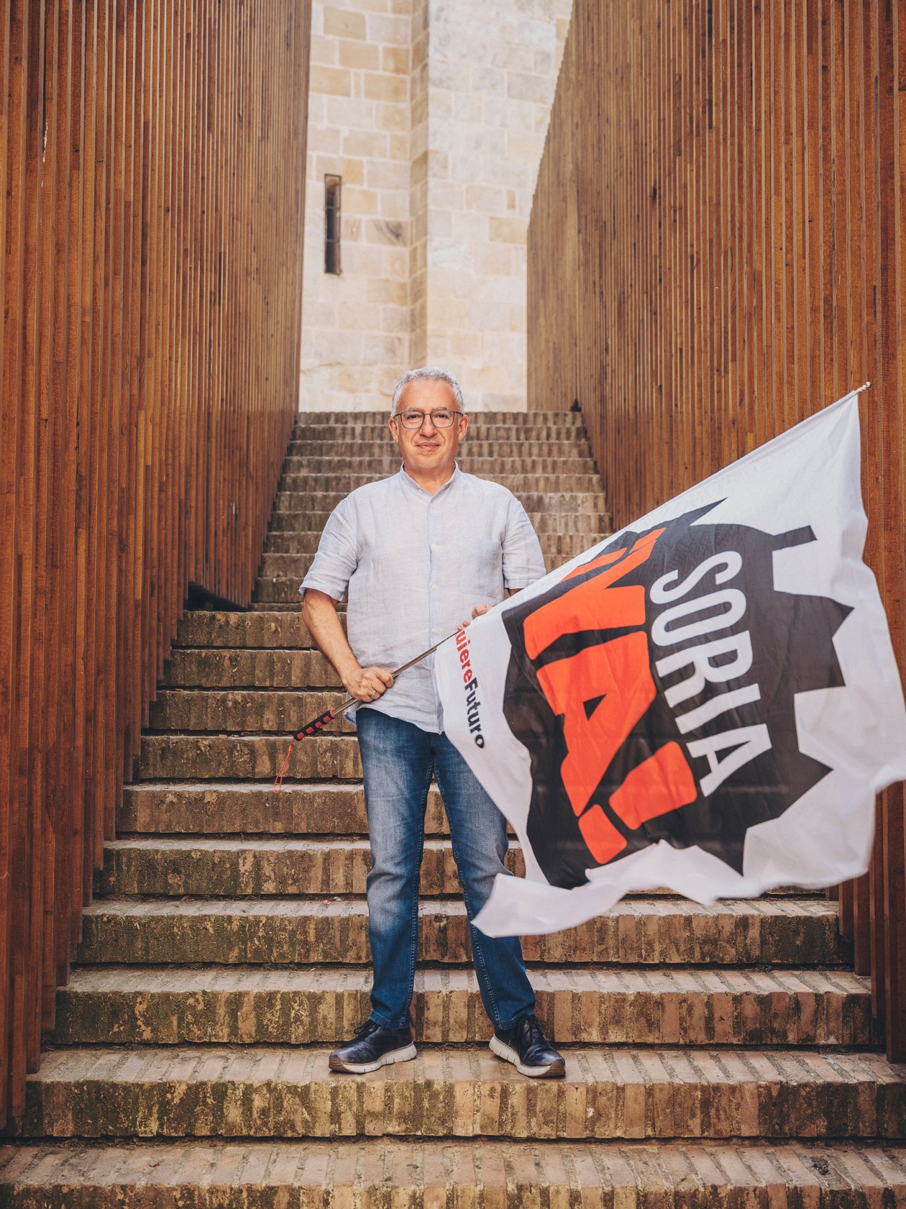 Ángel Ceña of Soria ¡Ya! holding a large flag with red and black logo while standing on stone steps