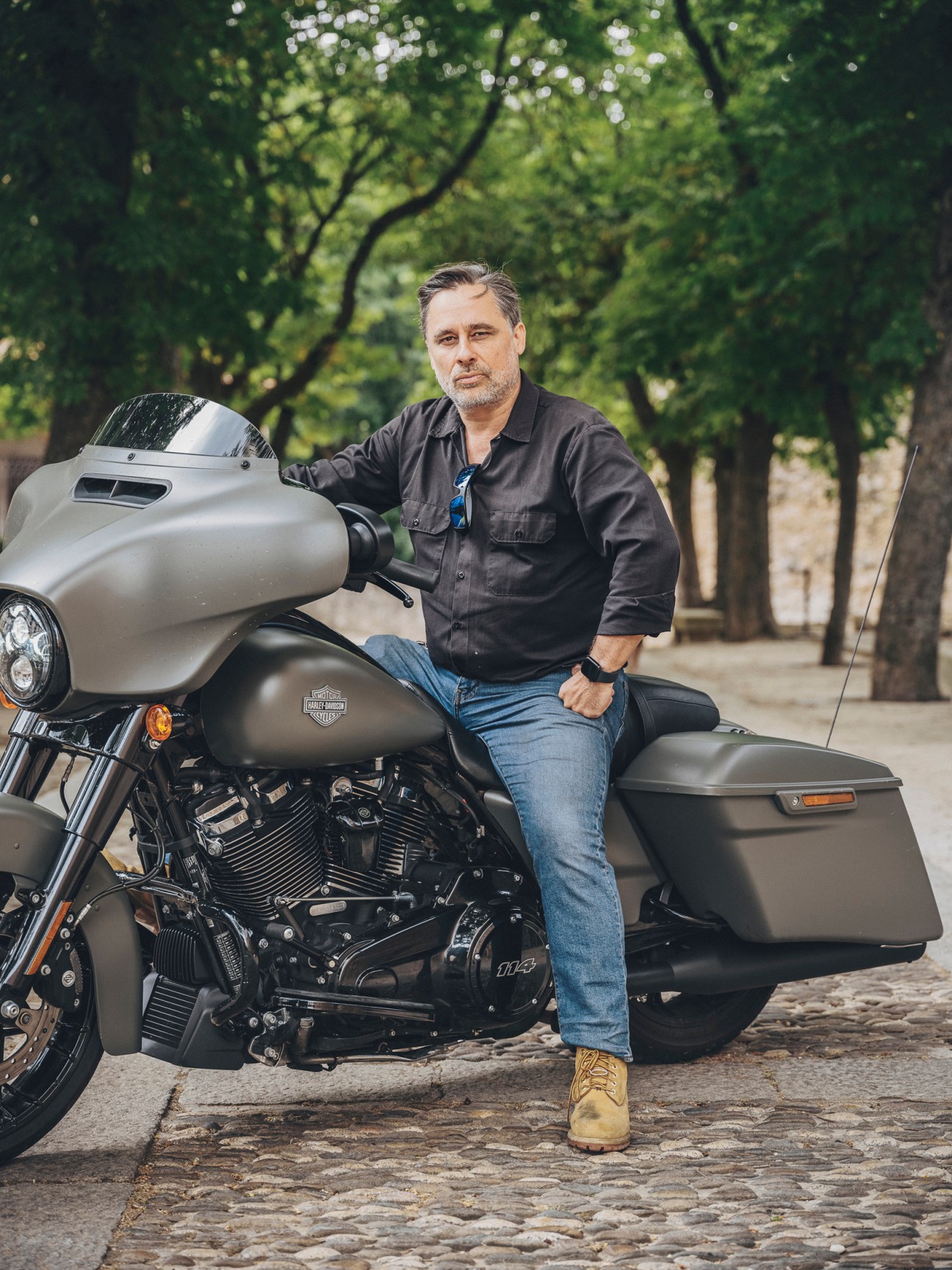 Ramón Pradera sitting on a Harley-Davidson motorcycle on a cobblestone street with trees in the background