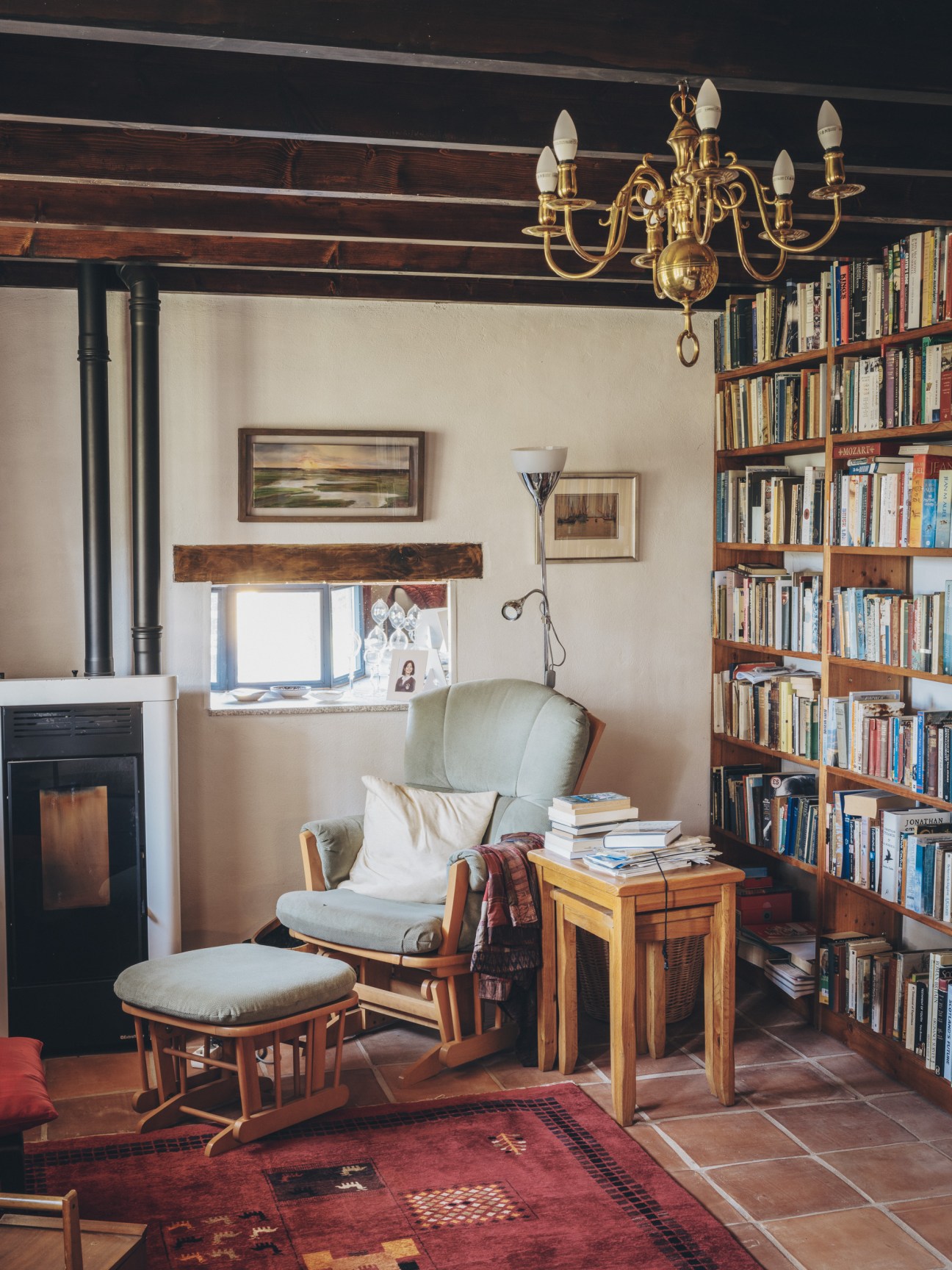 Cozy reading corner with light green armchair, ottoman, floor lamp, bookshelf, brass chandelier and exposed ceiling beams