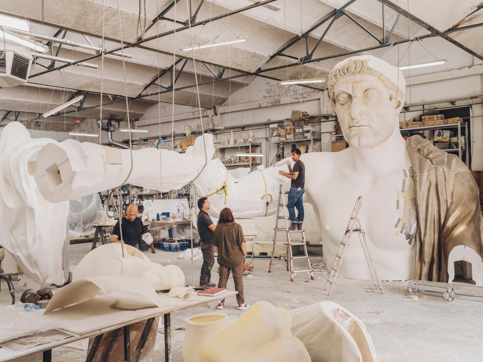 Workers applying finishing touches to large white statue parts in industrial workshop with scaffolding