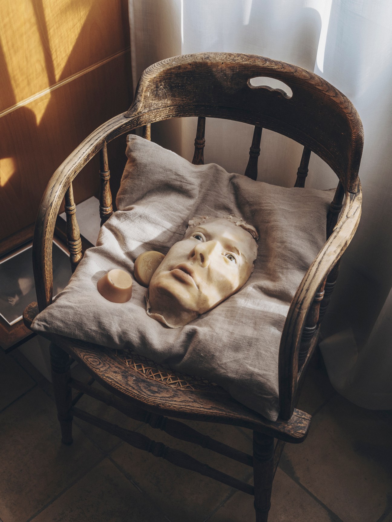 A broken sculptural face or mask resting on fabric in an ornate wooden chair by a window