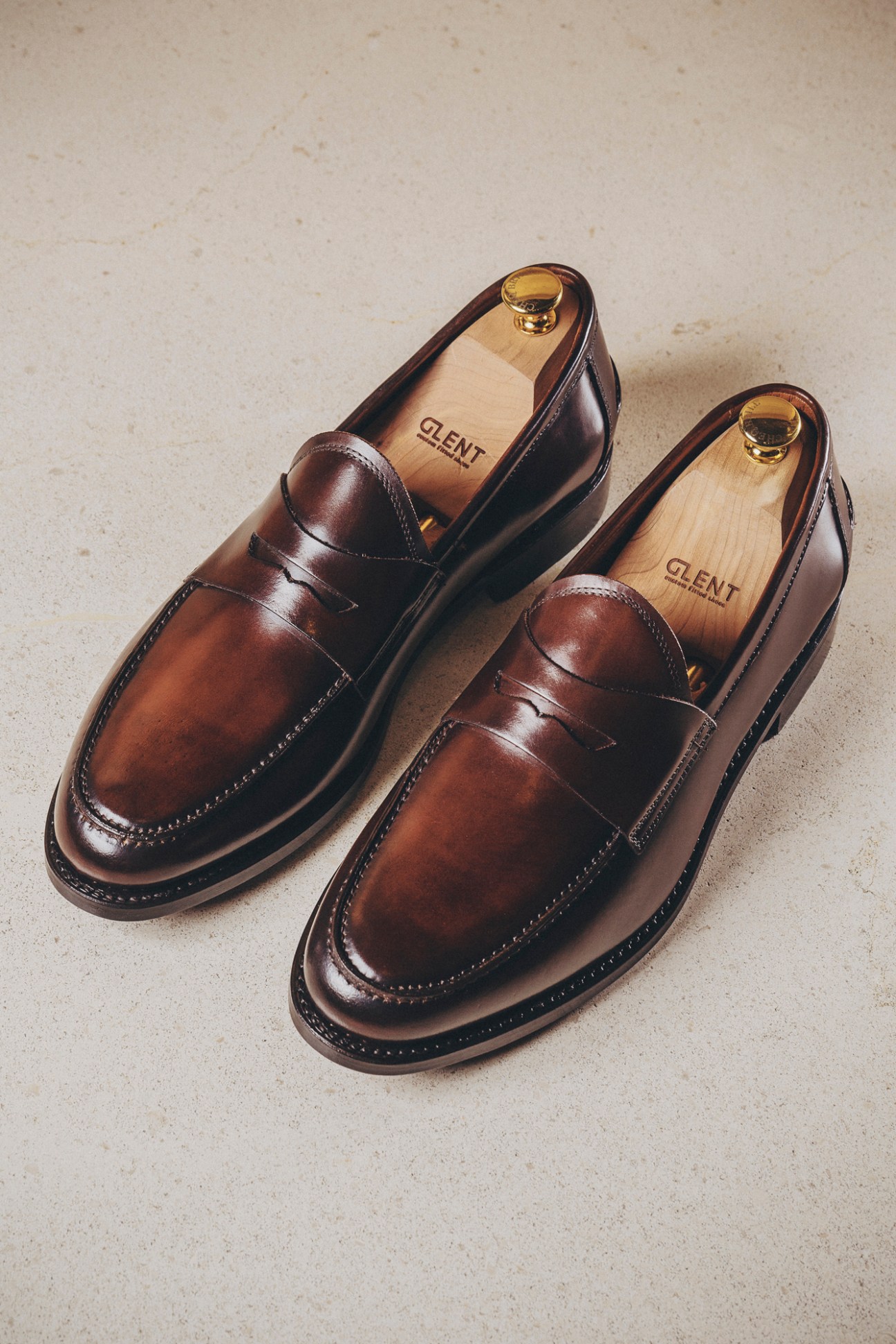 Brown leather penny loafers with wooden shoe trees and Glent branding on a light surface