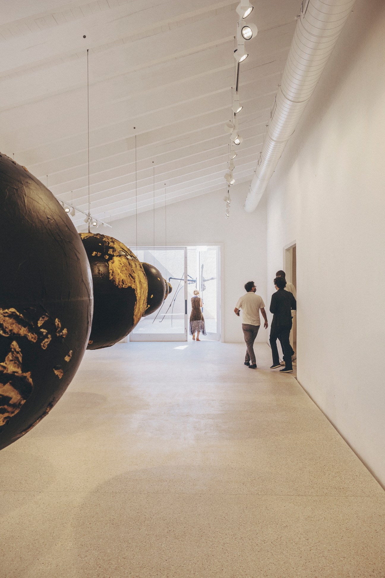 Two people walking through a modern white gallery space with large spherical artworks and track lighting