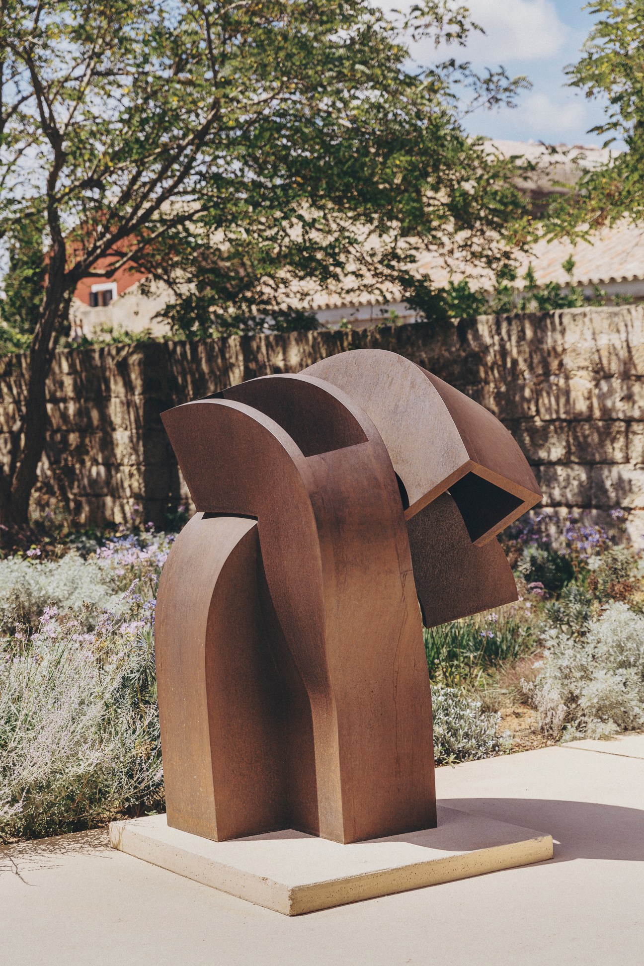 Large abstract metal sculpture with curved geometric forms in rust-colored patina displayed outdoors on stone base