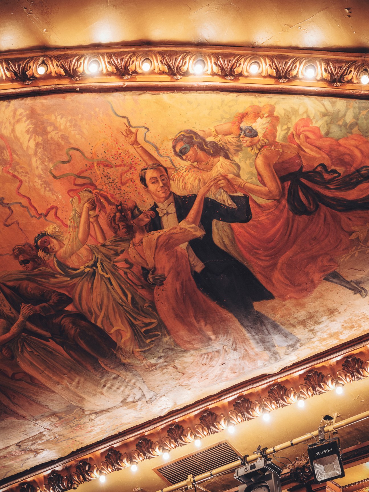 Ornate painted ceiling depicting aristocratic figures in flowing robes dancing, framed by decorative golden molding with lights