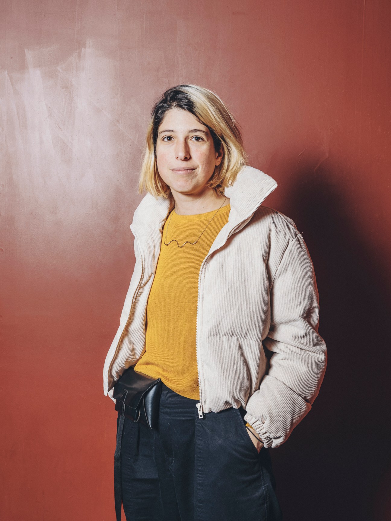 Woman in yellow top and beige jacket standing casually against red backdrop