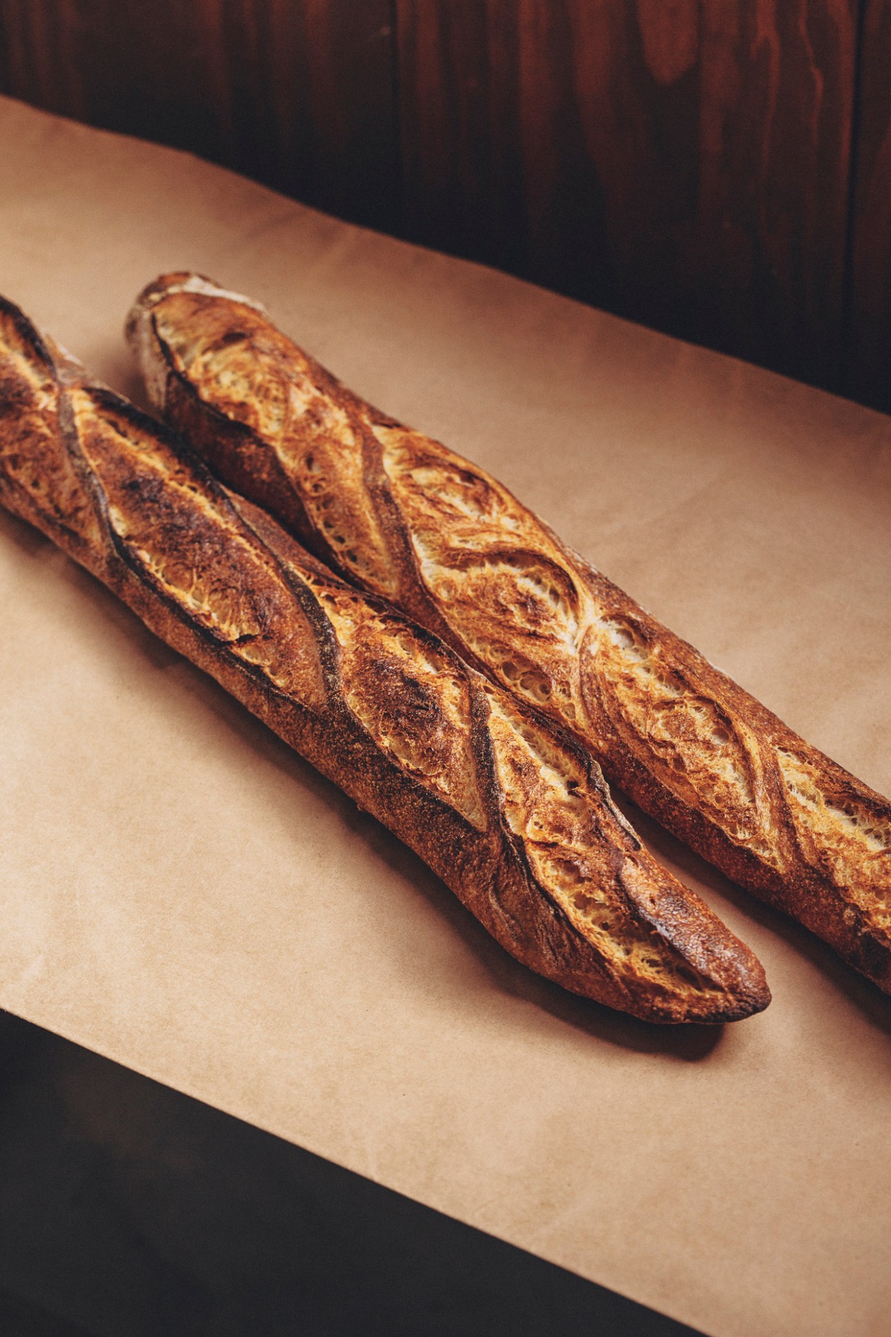 Three golden-brown pistola bread loaves wrapped in brown paper on wooden surface