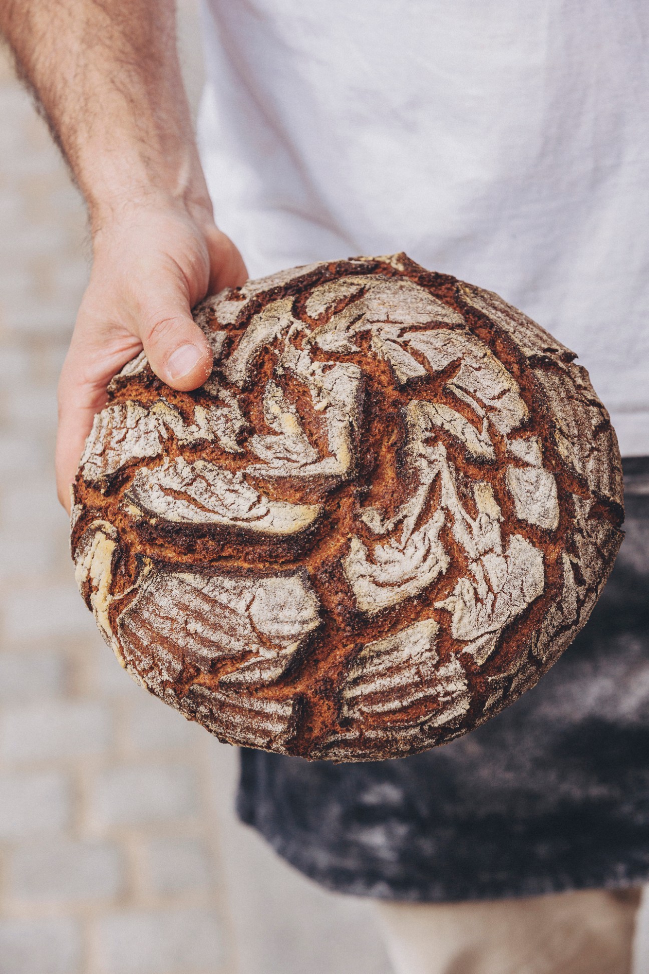 Hands holding a rustic dark rye sourdough bread loaf with decorative scoring patterns on the crust