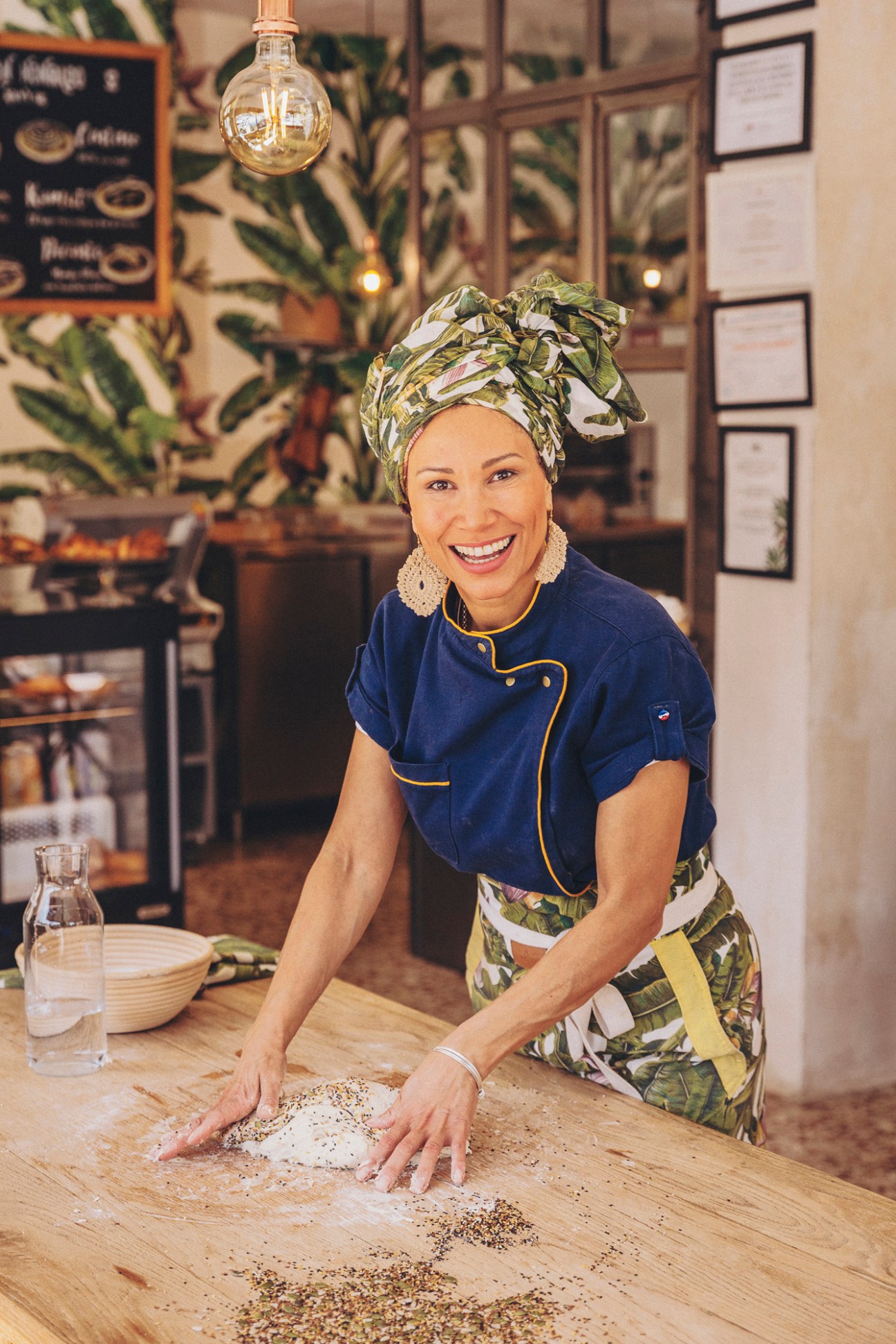 Woman in blue chef's jacket and colorful headwrap kneading dough on wooden counter at Amasa bakery