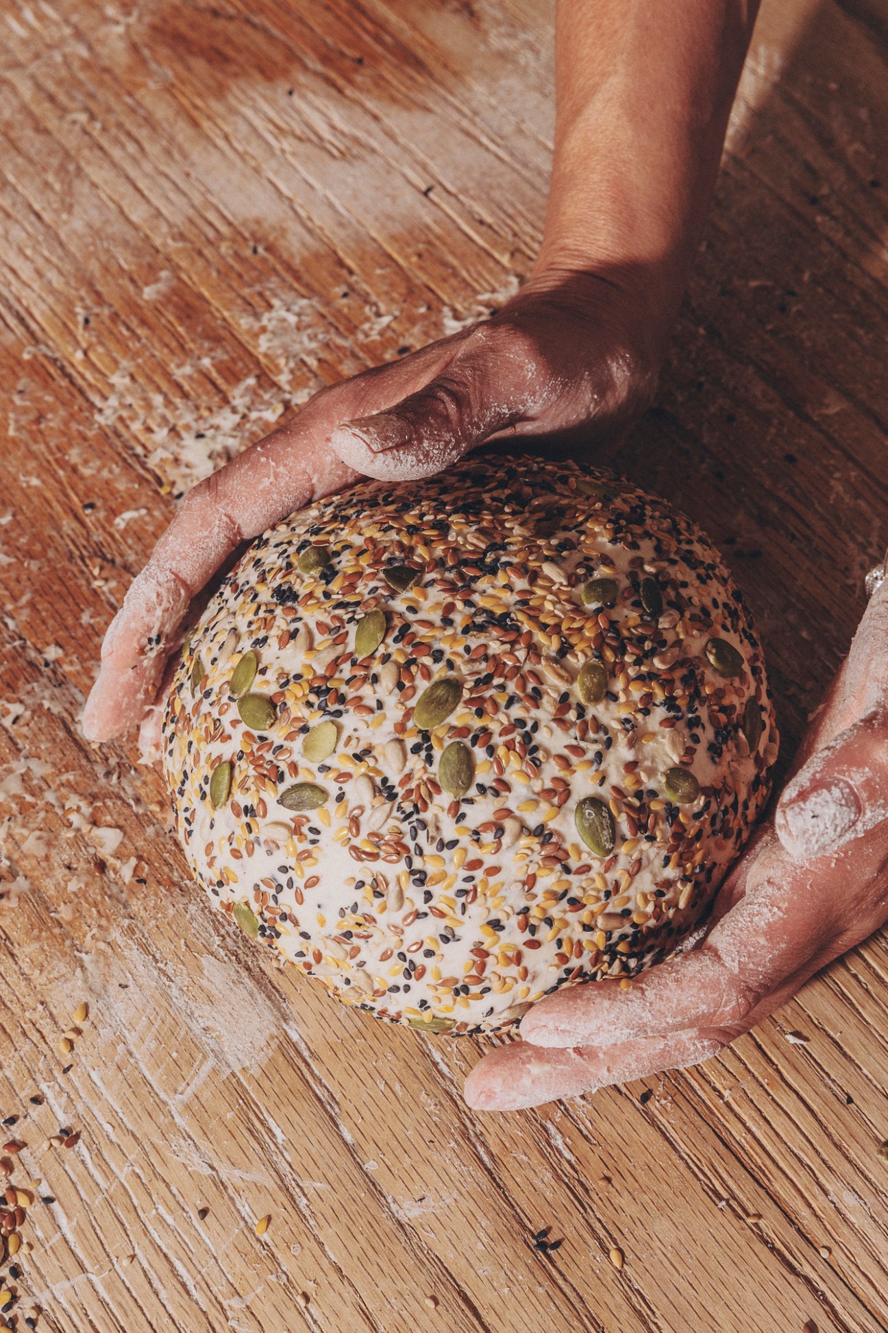 Close-up of hands protectively cupping a round seeded bread loaf covered in various seeds and grains