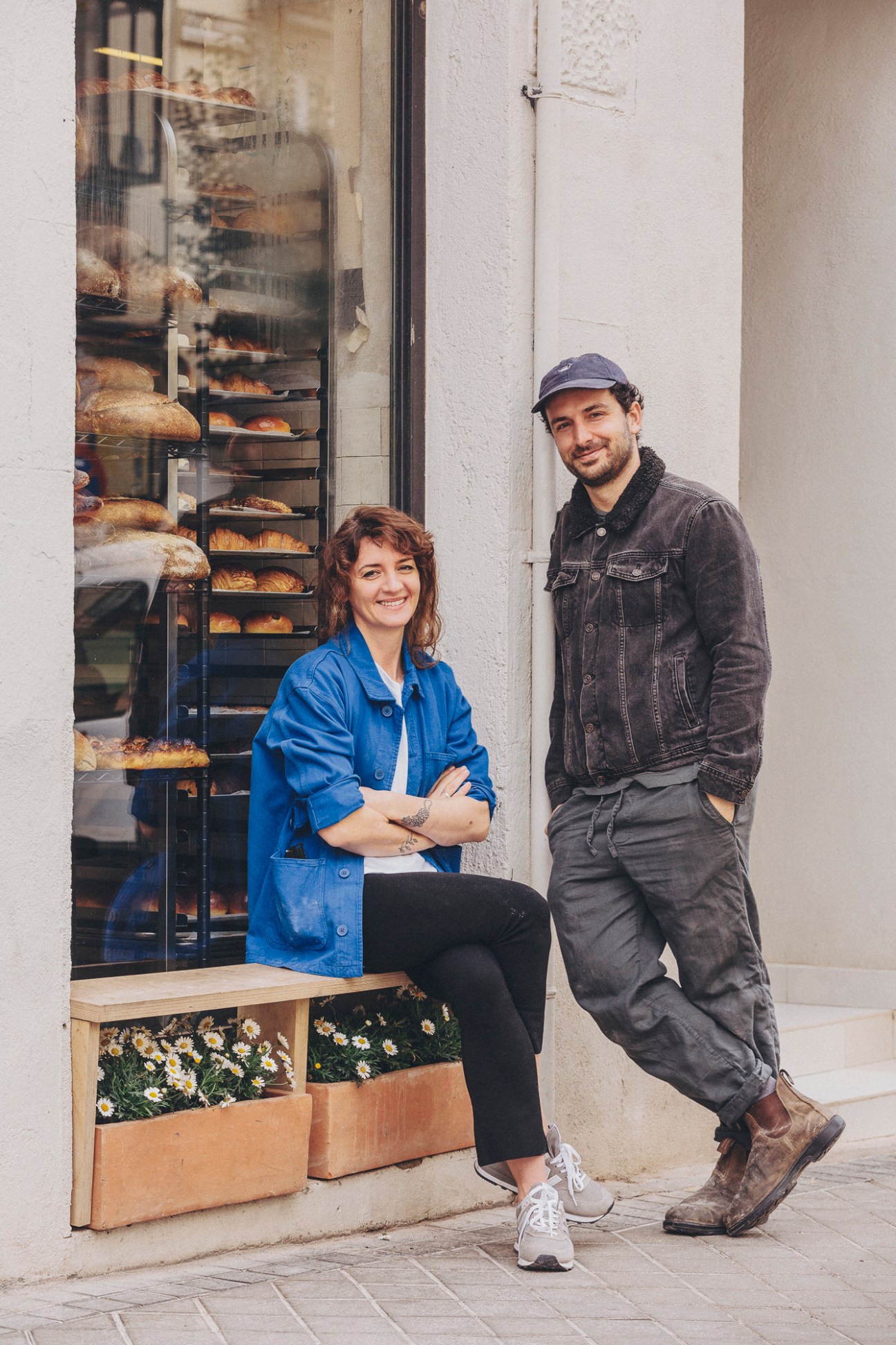 Two people standing outside Alma Nomad bakery, woman in blue jacket sitting on bench, man in denim jacket standing