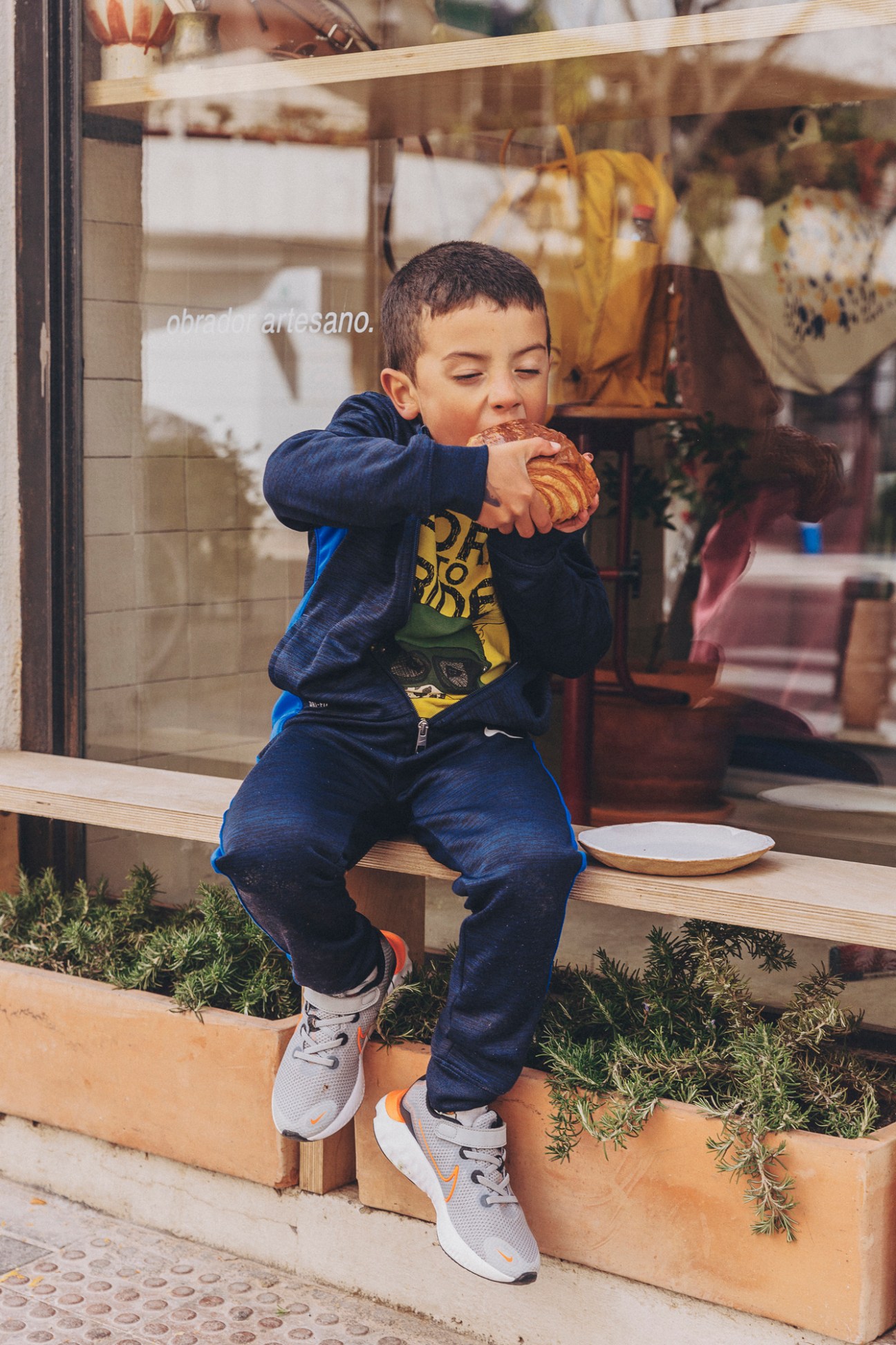 Young boy in navy blue jacket eating a pastry while sitting on wooden bench outside bakery