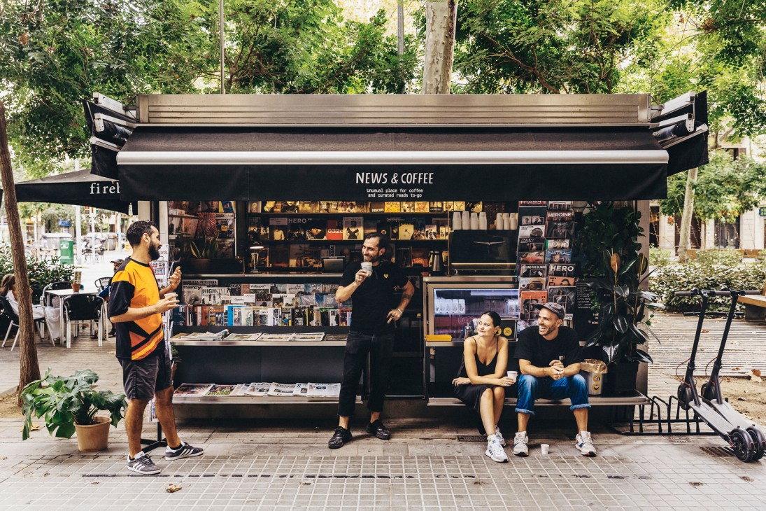Street newsstand kiosk with people sitting at bench, magazines displayed, and coffee service in Barcelona