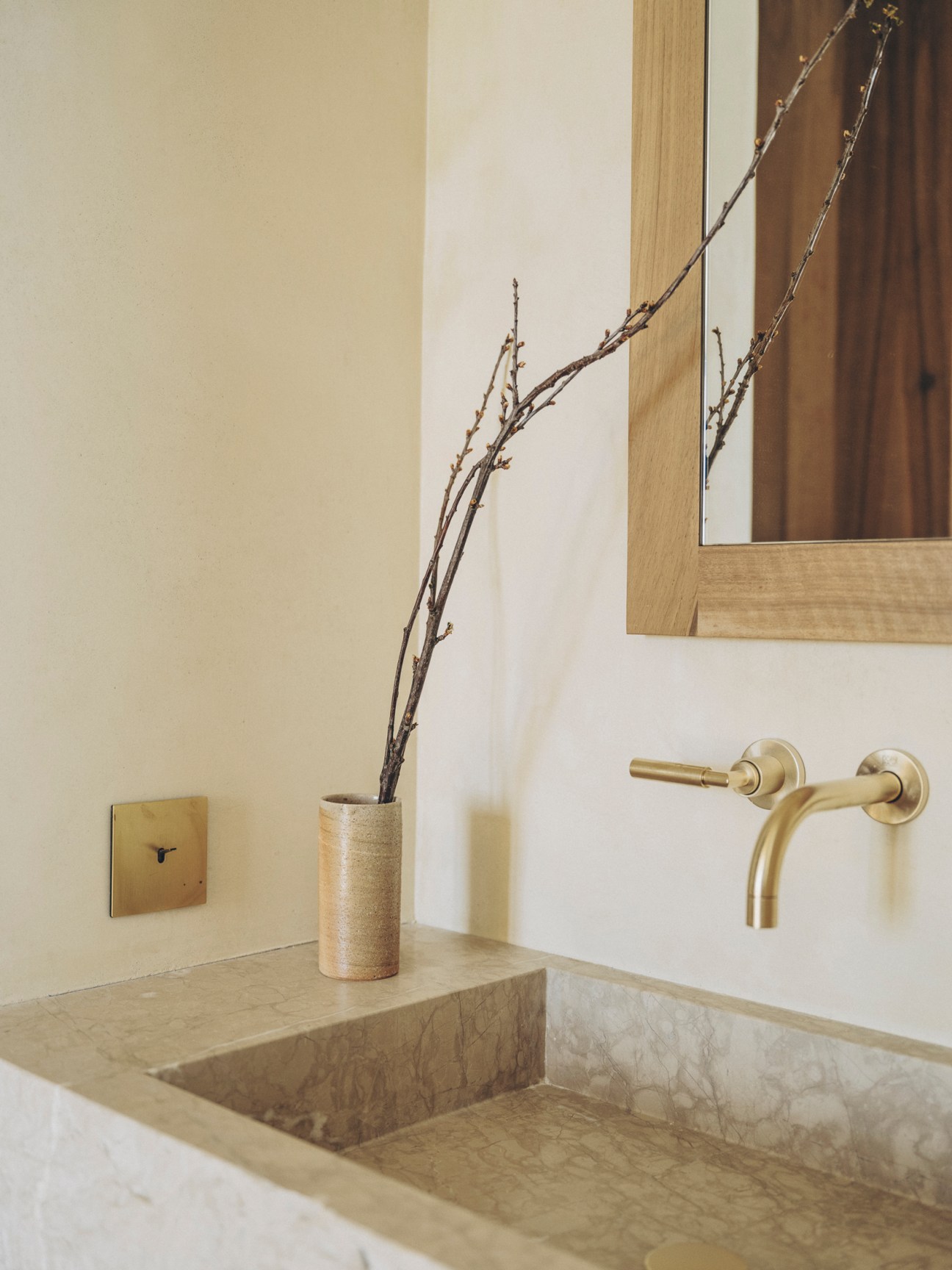 Stone washbasin with brass wall-mounted faucet and wooden vase containing branches