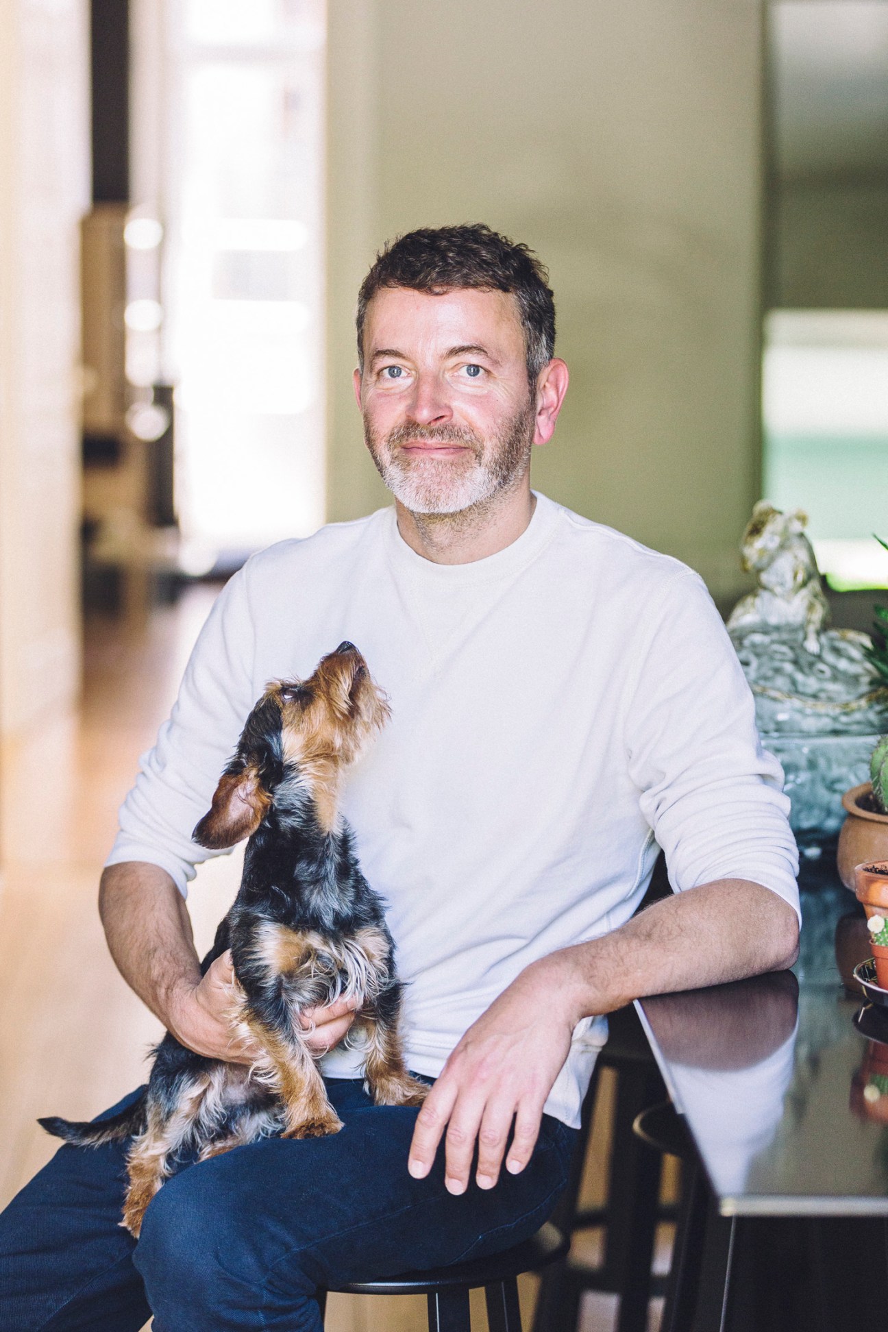 Man in white long-sleeved shirt holding a small black and tan dog indoors.