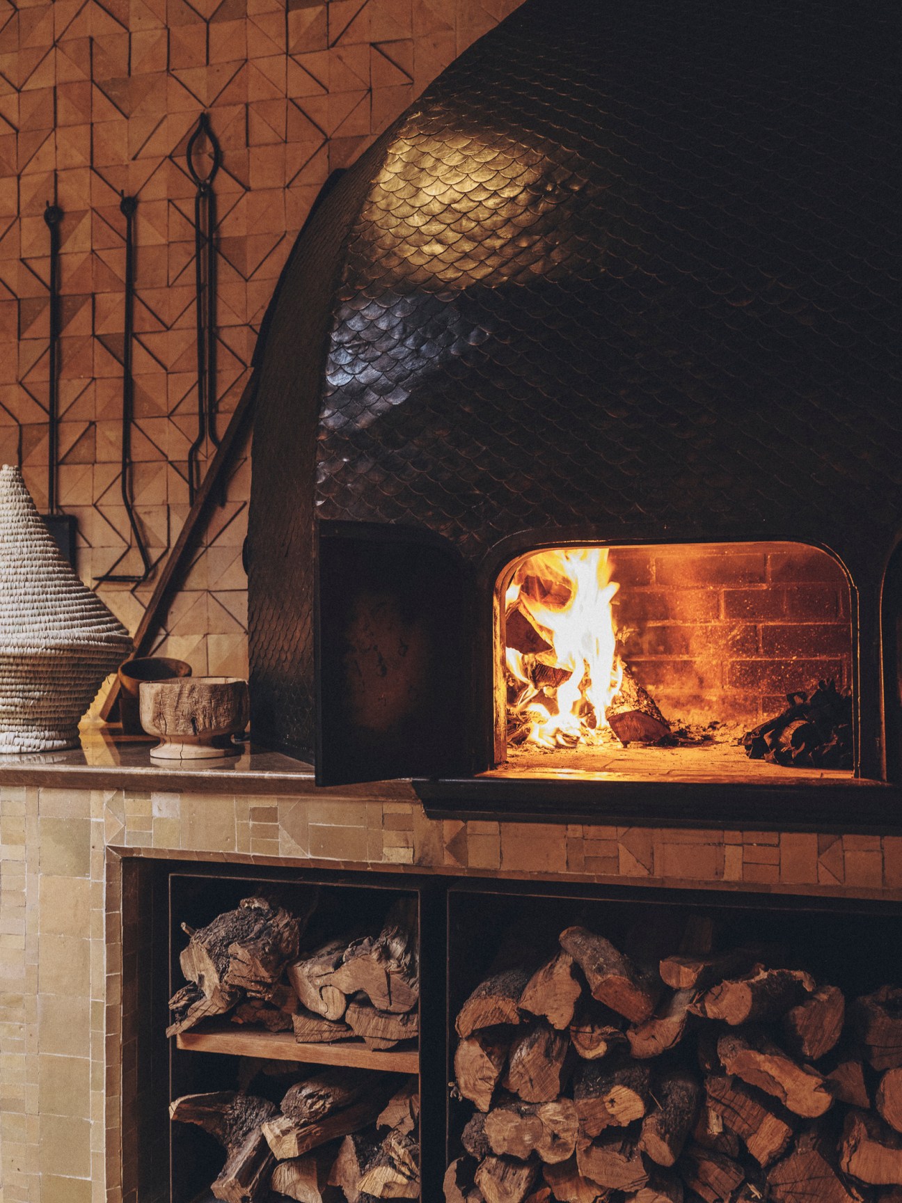 Wood-fired oven with glowing flames and stacked firewood storage below in restaurant kitchen