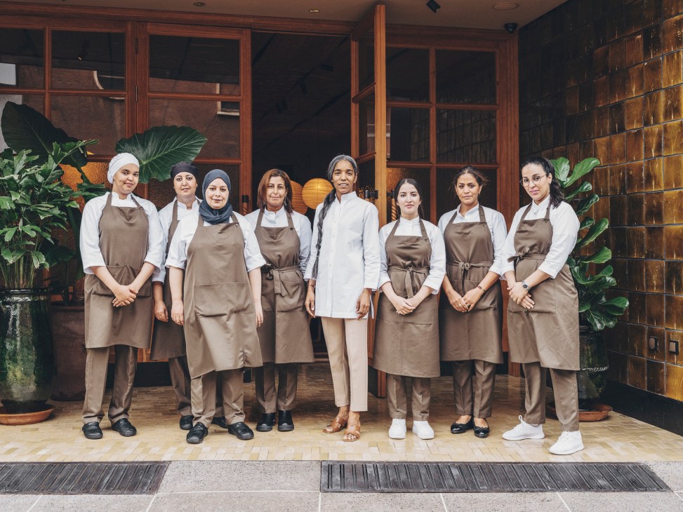 Eight women in brown aprons and white uniforms standing together in restaurant kitchen