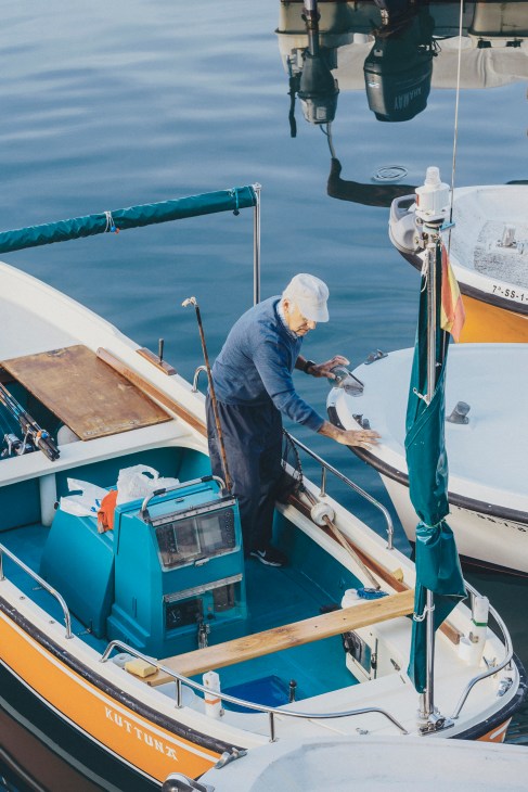 Fisherman in white cap working on yellow and blue fishing boat with nets and equipment visible.