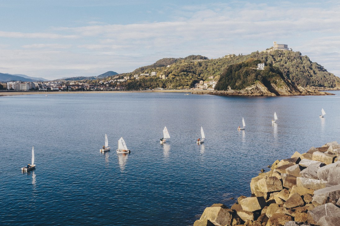 Sailboats on the Bay of Biscay with San Sebastián's coastal cityscape and forested headland in background.