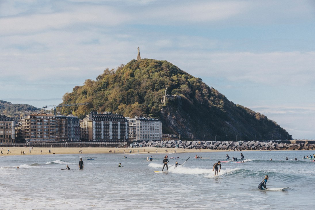 San Sebastián's shell-shaped La Concha beach with surfers in the water and a forested hill with a monument in the background.