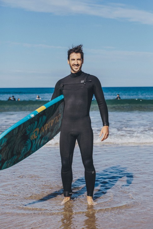 Man in black wetsuit holding surfboard on San Sebastián beach with ocean in background.