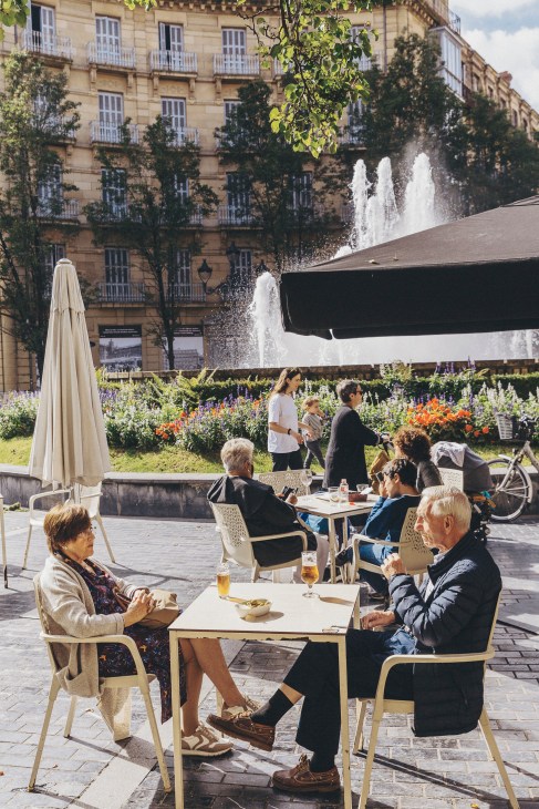 People dining and socializing at outdoor waterfront café tables with historic buildings and fountains in background.
