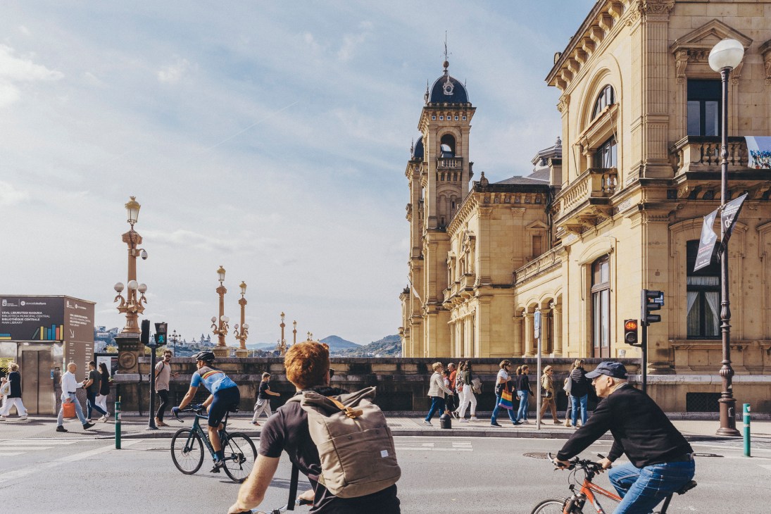 Cyclists and pedestrians on a sunny plaza in San Sebastián, Spain with historic buildings and mountains in background.