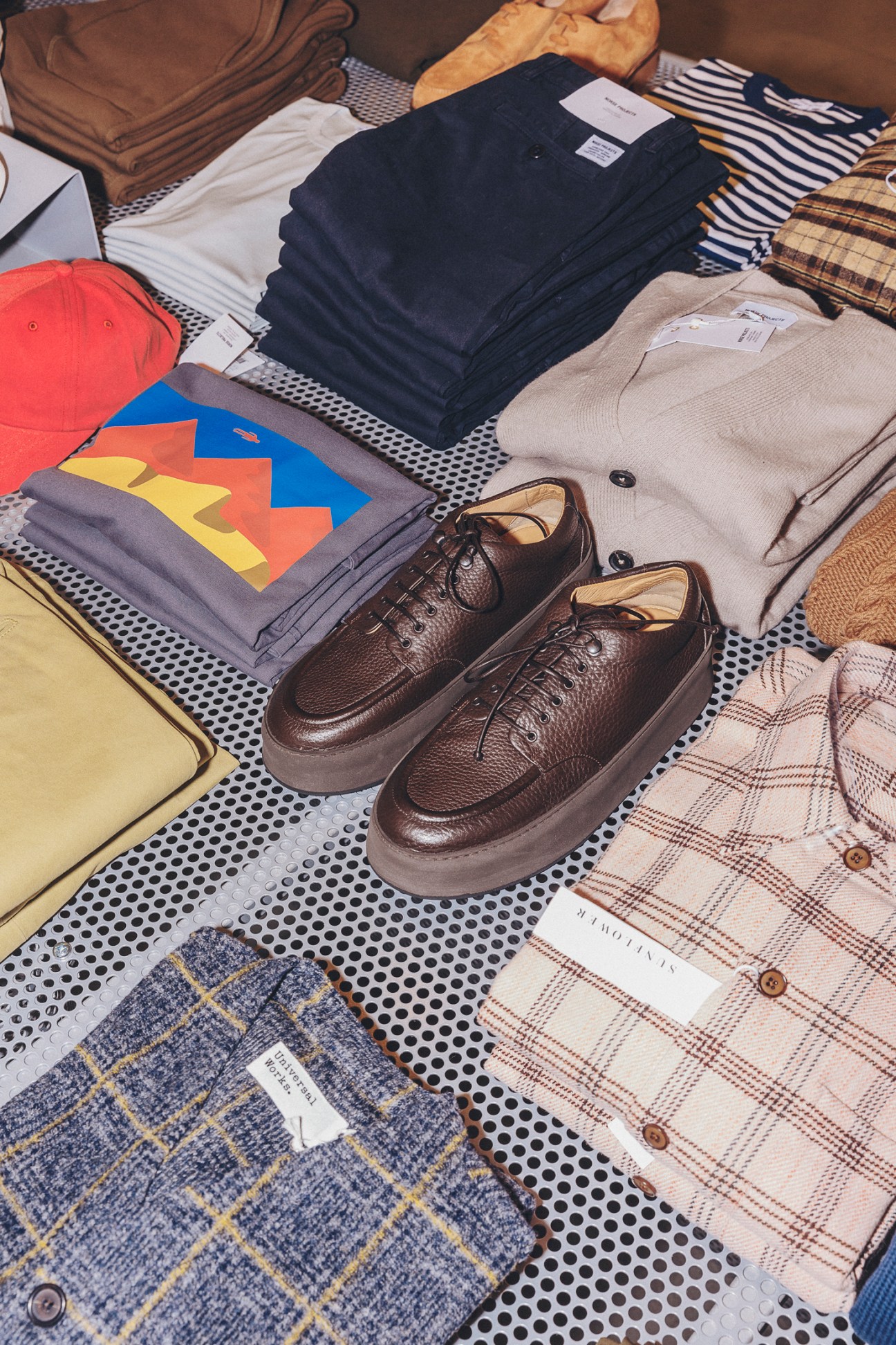 Flat lay of menswear items including brown leather shoes, folded clothing, and accessories on dotted surface