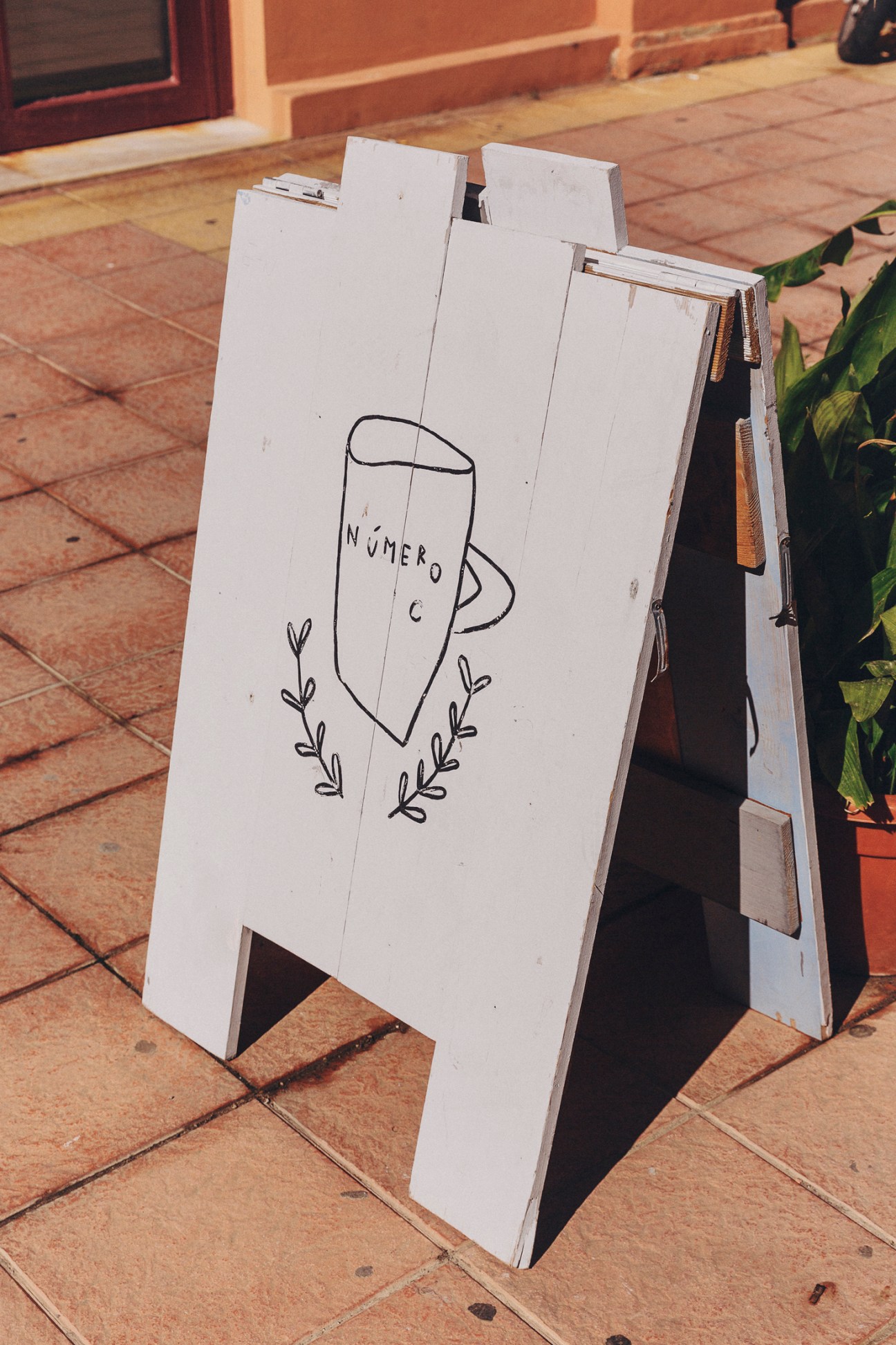 White A-frame sandwich board with hand-drawn illustration of Número C café logo and decorative leaf details on pavement.