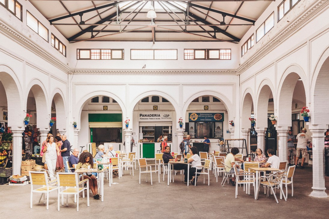 Interior of Tarifa's central market with white arches, wooden chairs, and shop fronts.