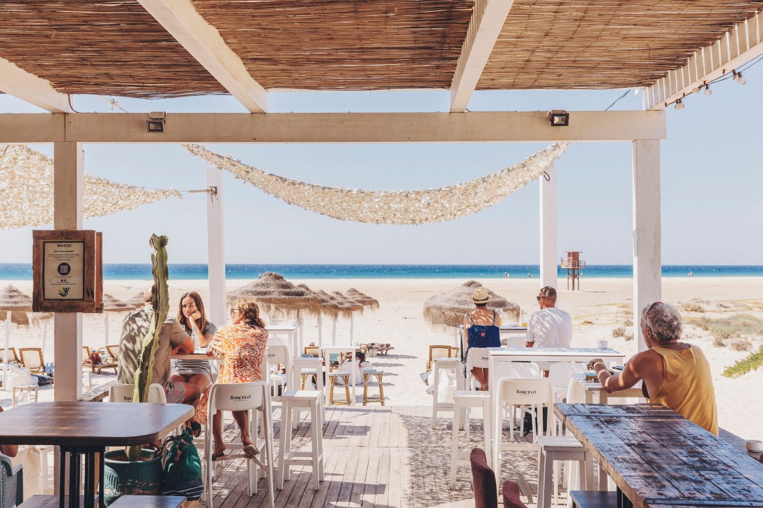 Beachgoers relax under a wooden pergola with straw shade coverings at the Waikiki chiringuito, overlooking the sand and Mediterranean Sea.