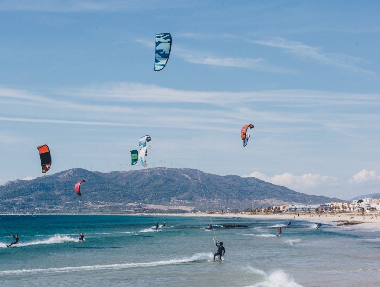 Multiple colorful kites flying above kitesurfers on Playa de los Lances beach in Tarifa with mountains in background