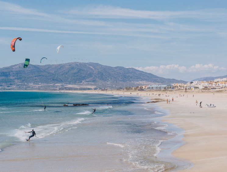 Multiple kitesurfers with colorful kites on Playa de Los Lances beach in Tarifa, Spain, with mountains and coastal town in background.