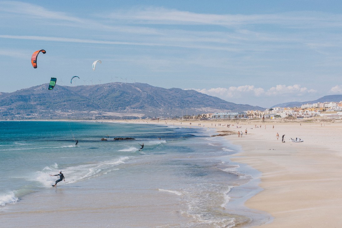 Multiple kitesurfers with colorful kites on Playa de Los Lances beach in Tarifa, Spain, with mountains and coastal town in background.
