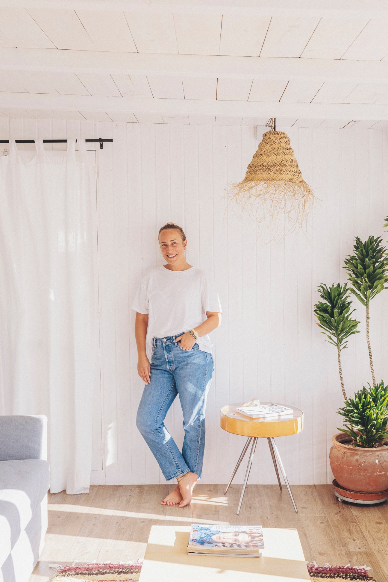 Fashion PR agent Eszter Felkai standing in a bright white room with woven pendant light and potted plants.