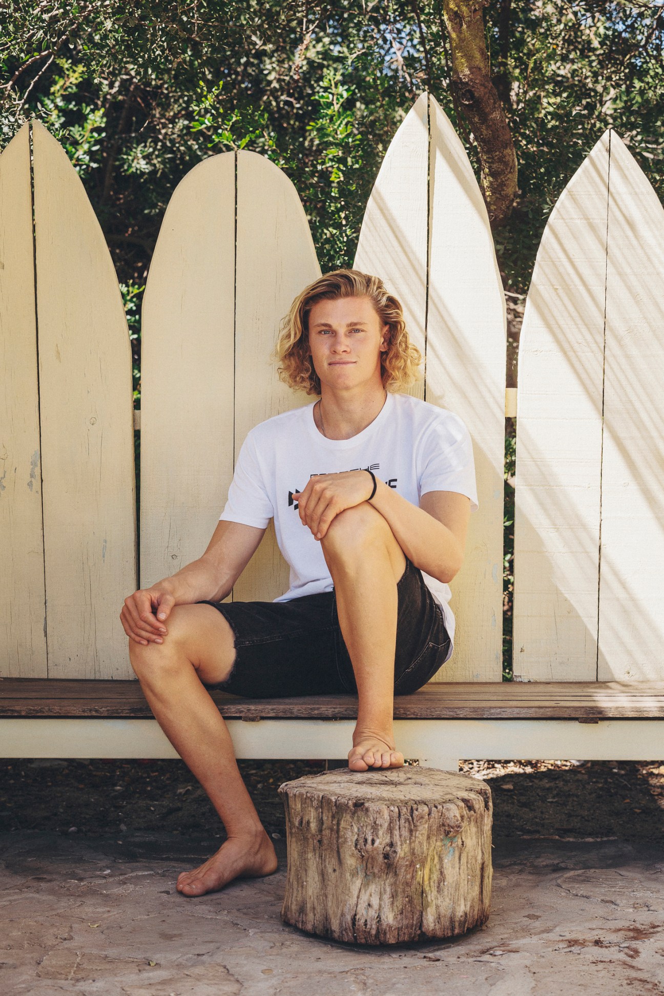 Professional kitesurfer Liam Whaley poses in front of white surfboards with ivy-covered trees behind him.